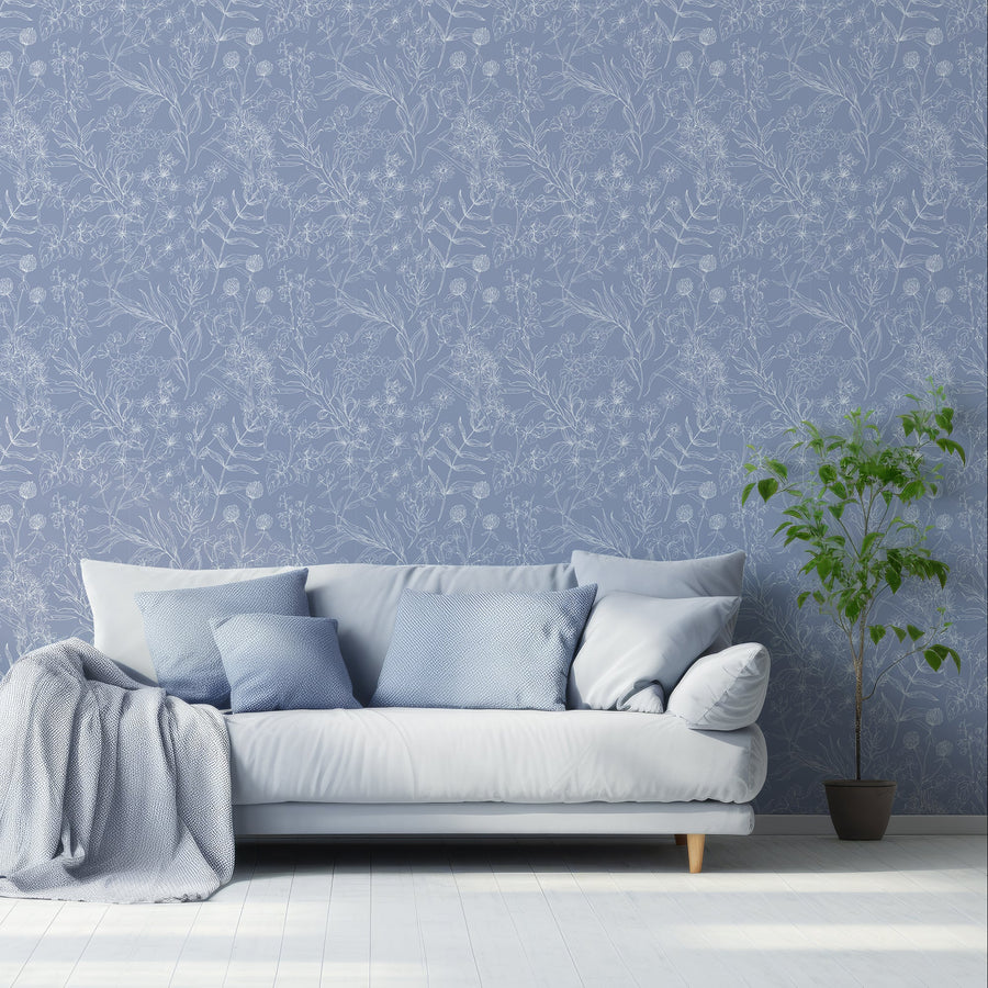 A living room with a baby bue fabric sofa with blue cushions and blanket next to a free standing plant, showing 'Estelle' which is a blue wallpaper featuring delicate botanical outlines of flowers, leaves and plants in light grey tones on a soft blue background in a repeating pattern.