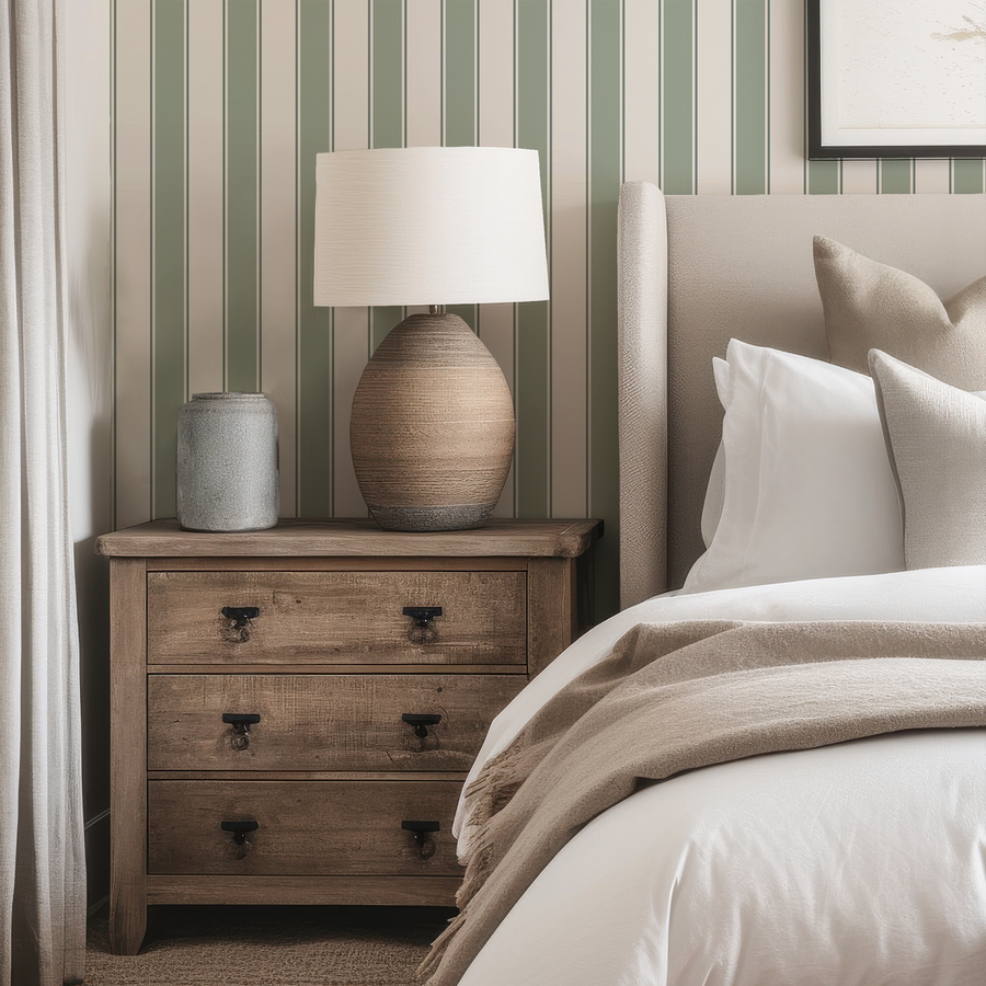 A neutral bedroom with a fabric bed frame and light coloured side table which has a vase and large lamp on it. The image features 'Nathalie' which is a classic stripe wallpaper featuring evenly spaced vertical lines in a simple repeating pattern with a clean, timeless design.