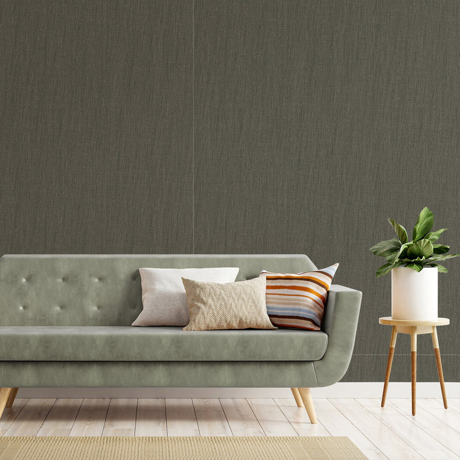 A living room with a modern sage green sofa with a side table next to it holding a plant, showing 'Hazel' which is a dark green wallpaper featuring a subtle fabric-effect texture with a woven pattern and grey undertones for a soft, sophisticated finish.