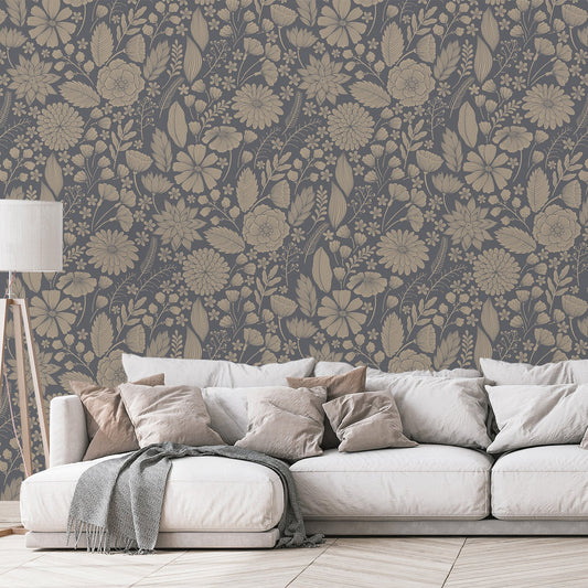 A living room scene with a beige sofa scattered with multiple cushions and a lamp showing 'Jessamine' which is a Blue botanical wallpaper featuring delicate floral blooms, leafy plants and fine botanical illustrations on a dark blue-grey background in a repeating pattern.