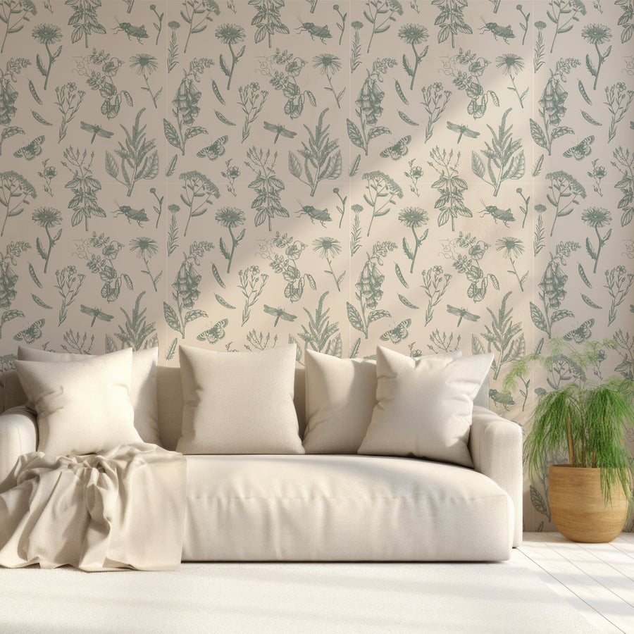 A living room with a cream sofa scattered with cushions next to a small green plant showing 'Lillian' which is a green botanical wallpaper featuring delicate foxglove flowers, white daisies, leafy plants and butterflies on a soft cream background in a vintage-inspired repeating pattern.