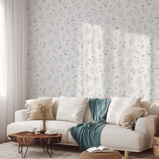 A living room with a cream sofa scattered with pillows and a blue blanket showing 'Nola' which is a soft vintage wallpaper featuring delicate wildflower line drawings in pastel green tones on a gentle grey background in a repeating botanical pattern.