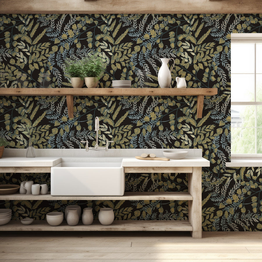 A cottage themed kitchen with light wooden fittings featuring Ottilie wallpaper which is vintage-style golden leaves with green foliage on a deep black background in a repeating botanical pattern.