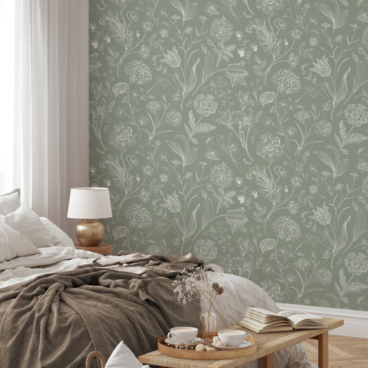 A relaxed styled bedroom showing 'Priscilla' which is a botanical wallpaper featuring delicate white floral and plant illustrations on a soft green background in a vintage-inspired repeating pattern.