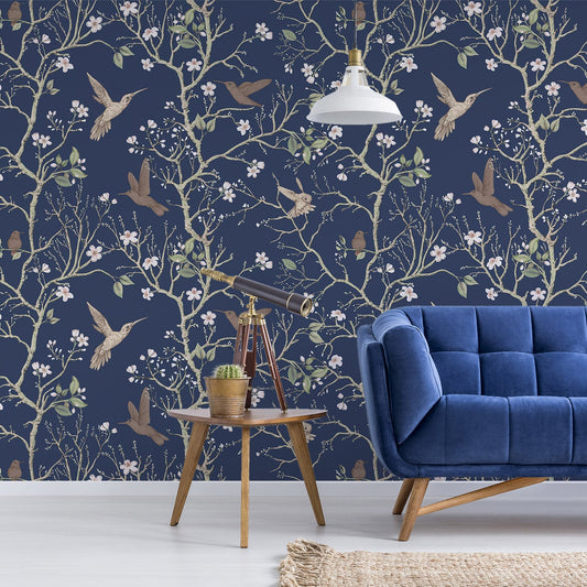 A living room with a blue tufted sofa and a small side table showing 'Denora' which is a dark navy wallpaper featuring hand-drawn chinoiserie trees, birds and colourful flowers in an elegant repeating botanical pattern.