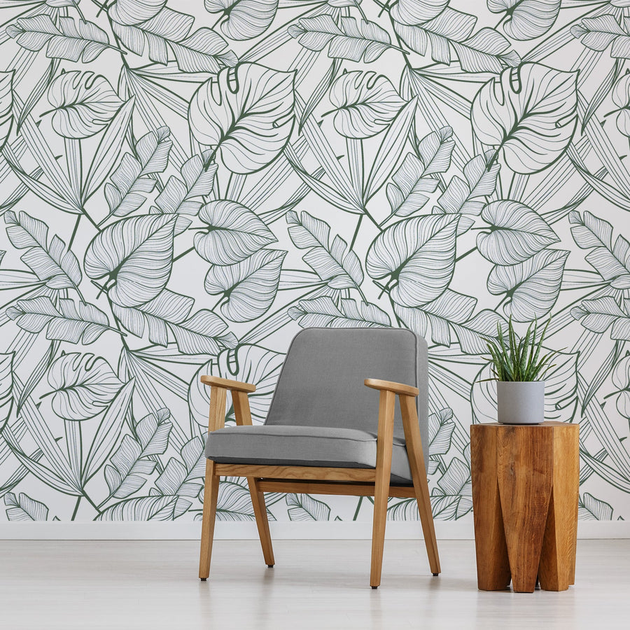 A living room with a grey chair with a small side table with a plant on top of it featuring 'Juniper Dark Green' which is tropical wallpaper featuring dark green outlined palm leaves and lush botanical foliage on a crisp white background in a bold repeating pattern.