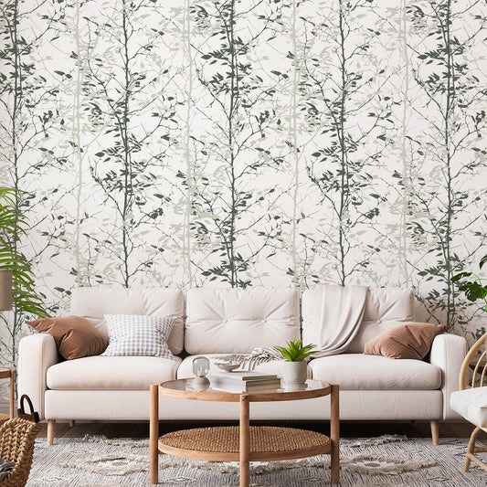 A living room with a cream sofa, scattered with cushions showing 'Kiri' which is a green wallpaper featuring layered forest trees in varying shades of green with a hand-painted botanical landscape design.