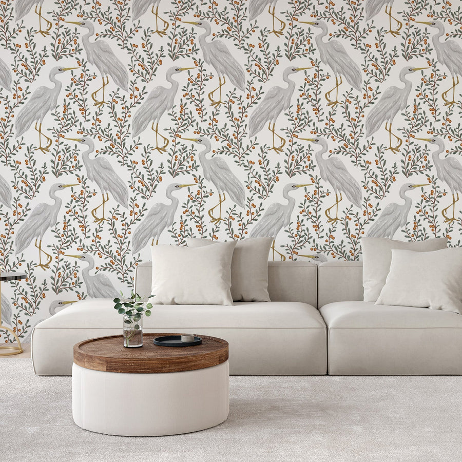 A living room with a white sofa and coffee table showing the wallpaper Tallulah which is a botanical wallpaper with illustrated herons, leafy branches and berries on a light background in a vintage-inspired repeating design.
